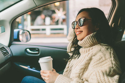 Side view of smiling woman with disposable cup sitting in car