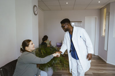 Smiling male doctor greeting female patient sitting in waiting room at clinic