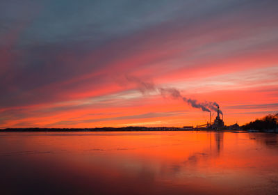Silhouette factory by sea against orange sky