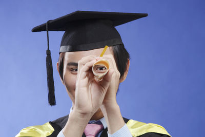 Portrait of man in graduation gown looking through certificate over blue background