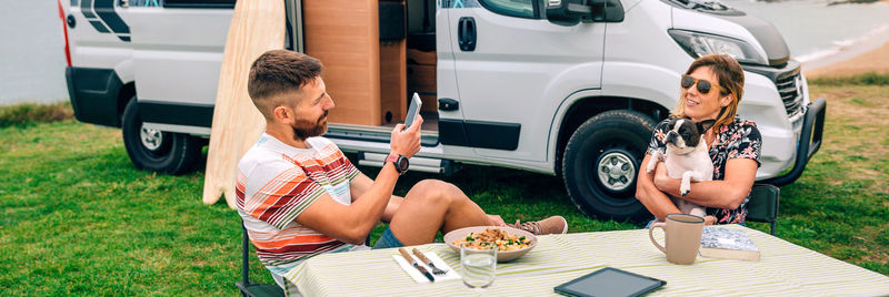 Man taking photo to woman hugging dog with van and beach on background