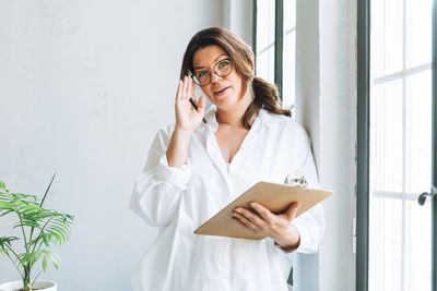 Portrait of young smiling brunette woman plus size near window with pen and documents in hands 