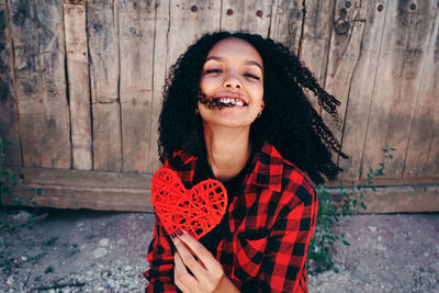 Portrait of cheerful woman holding red heart shape against wooden wall