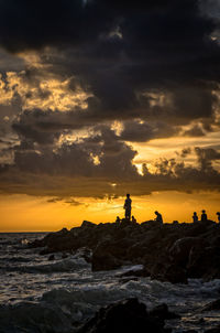 Silhouette people on rock formation in sea against cloudy sky during sunset