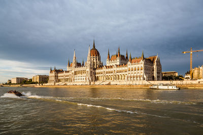 Hungarian parliament building by danube river against cloudy sky