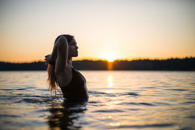 Girl in a long black swimsuit swims outside the city on the lake in the rays of sunset or dawn.