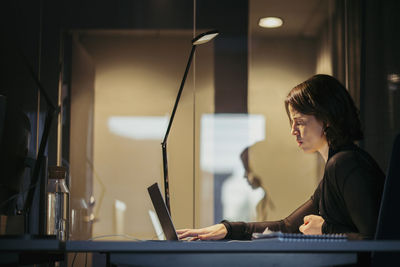 Side view of businesswoman using laptop at desk in dark office