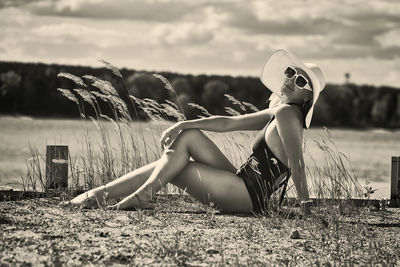 A woman in a swimsuit, hat and sunglasses sunbathes in summer on the riverbank among the grass