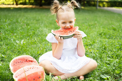Funny smiling kid girl in white dress eating watermelon at green lawn