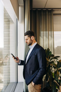 Side view of businessman using smart phone while standing near window in coworking office