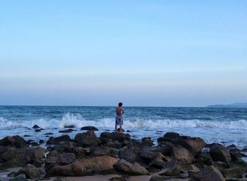 Shirtless man standing on rocks at sea shore against sky during sunset