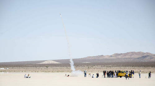 People looking at rocket while standing on sand against clear sky