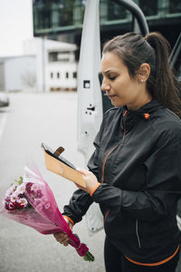 Serious delivery woman looking at mobile phone while holding bouquet of flowers in city