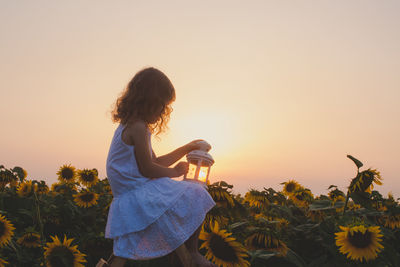 Rear view of woman standing by sunflower against sky during sunset