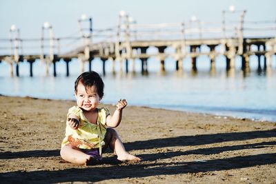 Full length of cute baby girl sitting at beach against bridge