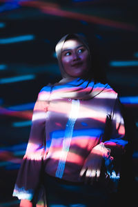 Woman looking away while standing in dark room with multi colored lights