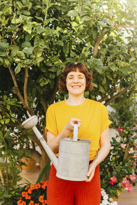 Smiling woman with watering can standing in front of lemon tree at garden
