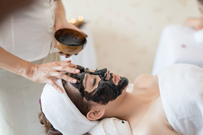 Beautician applying coal facial mask on face of young woman in spa