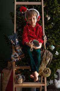 Barefoot boy in an elf costume holds craft paper deep green christmas tree ball