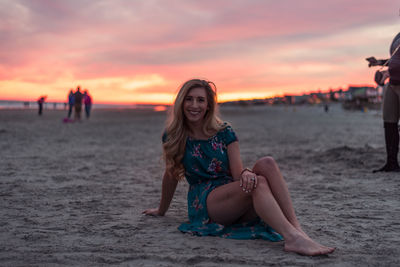 Young woman sitting at beach against sky during sunset