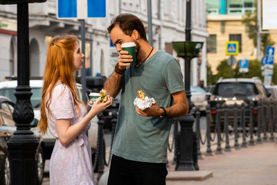 Young couple talking while walking along city street and eating takeaway food