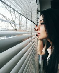 Thoughtful beautiful woman looking through window blinds