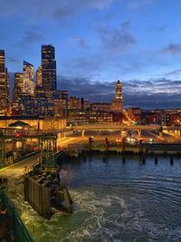 Illuminated buildings by river against cloudy sky
