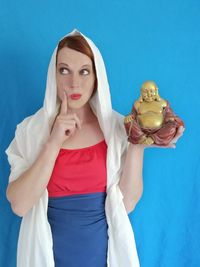 Thoughtful woman holding laughing buddha figurine against blue wall