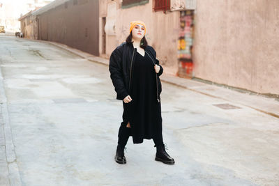 Young female with green dyed hair wearing a beanie and a black jacket looking at camera while standing on the street with buildings on sunny street