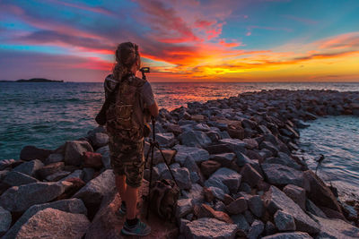 Man photographing while standing on rocks at beach against sky during sunset