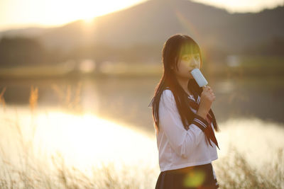 Portrait of young woman drinking water while standing by lake during sunset