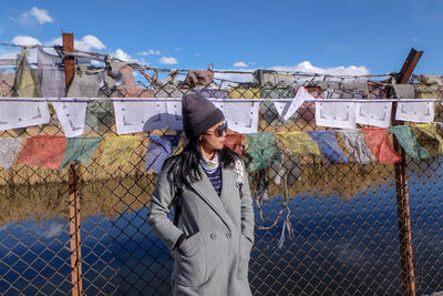 Woman wearing warm clothing while standing against fence and prayer flags