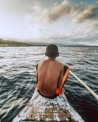 Rear view of shirtless boy sitting on boat in sea against sky