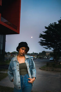 Portrait of young woman standing in city against sky at dusk