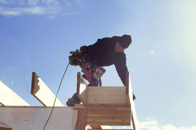 Low angle view of worker with drill standing on roof beam against sky