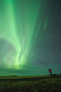 Remote view of unrecognizable traveling photographer taking picture of green aurora borealis shining in night sky in lofoten islands