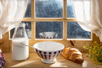 Close up of milk and croissant with bowl against window