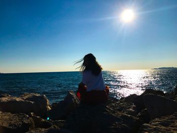 Rear view of woman sitting on rock by sea against clear blue sky