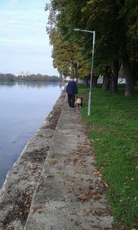 Rear view of grandfather with kid walking on footpath by lake against sky