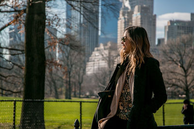 Happy woman wearing jacket standing by trees and buildings