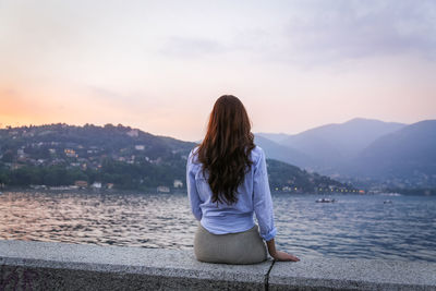 Rear view of woman sitting by sea against mountains during sunset