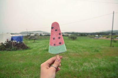 Cropped hand of woman holding ice cream over field against sky