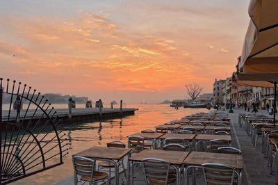 Tables and chairs at sidewalk cafe by sea during sunset