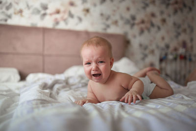 Charming baby, 6 months old, lies crying on his stomach on a bed in a bright, real interior,
