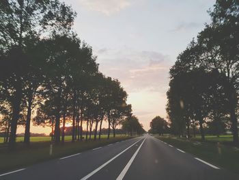 Empty road by trees against sky during sunset