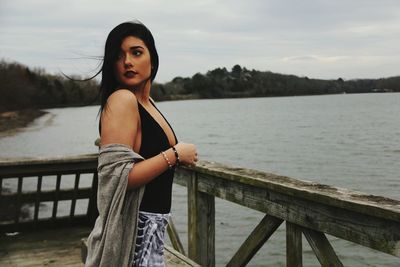 Portrait of young woman standing by railing against river