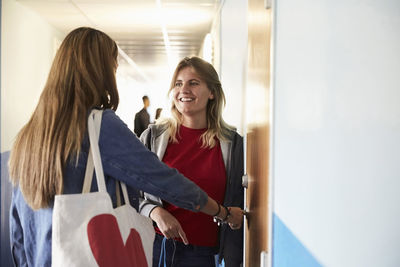 Smiling female friends talking while standing by door in corridor at university on sunny day