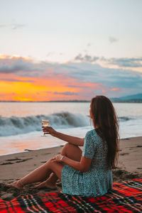 Woman sitting on beach against sky during sunset