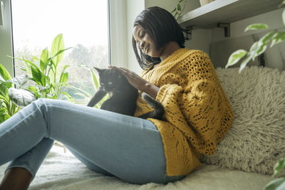 Smiling woman with cat sitting by window at home