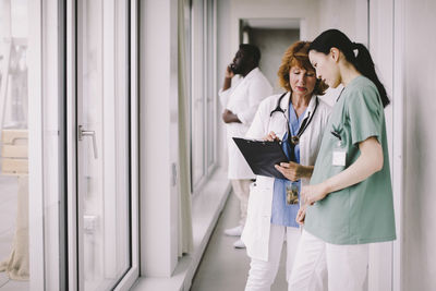 Senior female doctor discussing over record with nurse while standing in hospital corridor